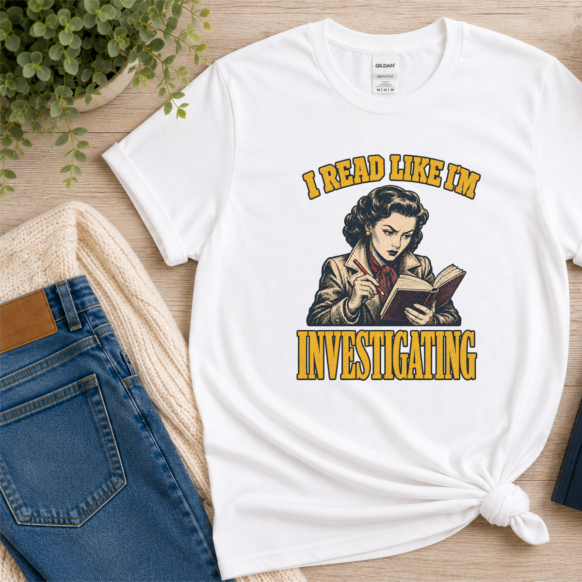 White T-shirt laid flat with an illustrated reader holding a book and pen and the words “I Read Like I’m Investigating.”