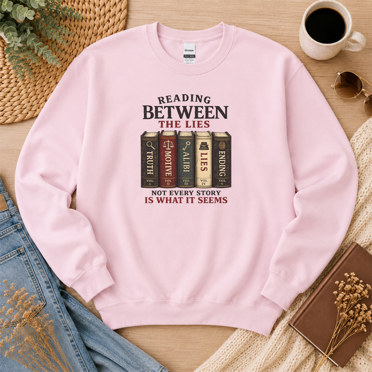 Light pink crewneck sweatshirt laid flat with books labeled Truth, Motive, Alibi, Lies and Ending and the words “Reading Between the Lies. Not Every Story Is What It Seems.”