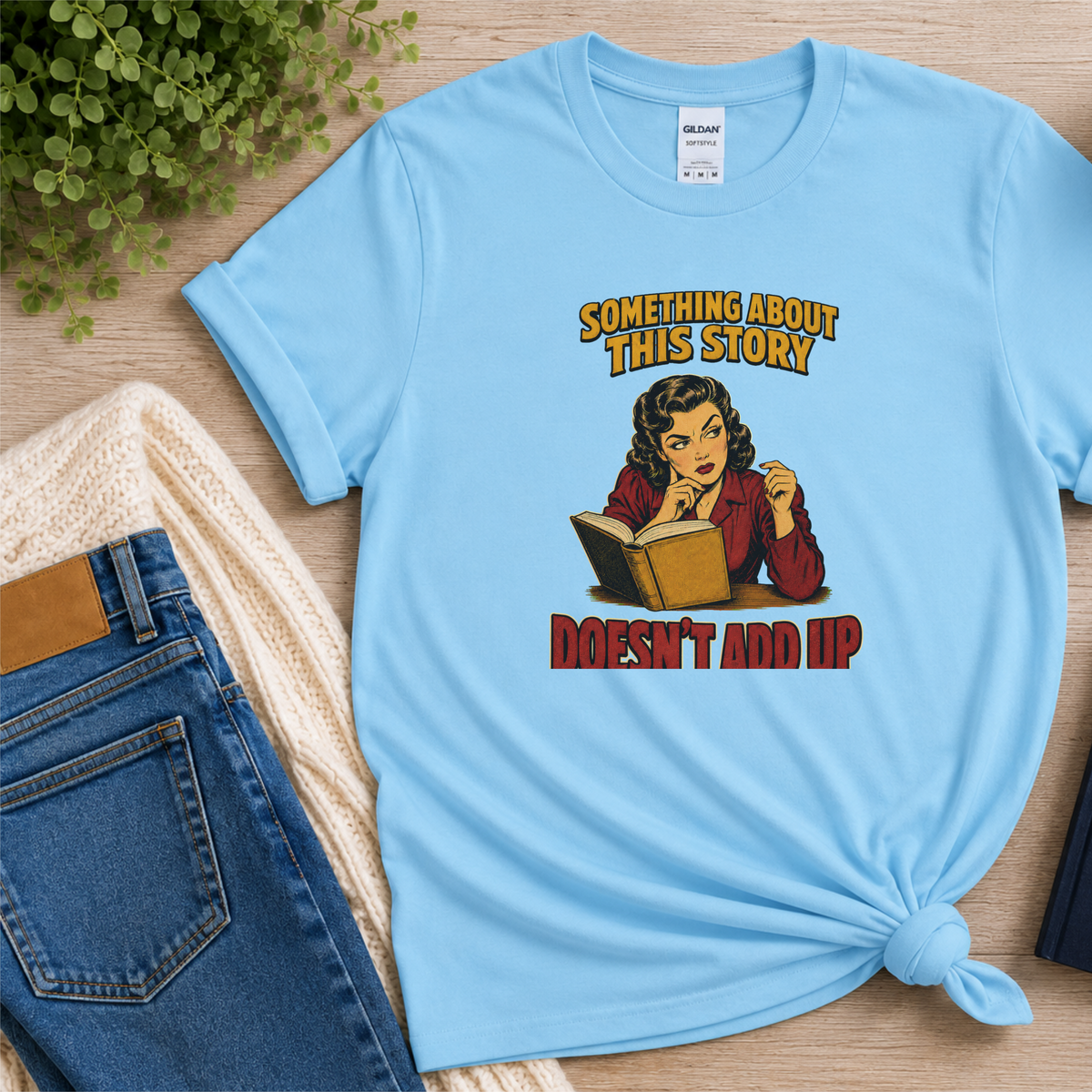 Light blue T-shirt laid flat with an illustrated reader holding an open book and the words “Something About This Story Doesn’t Add Up.”
