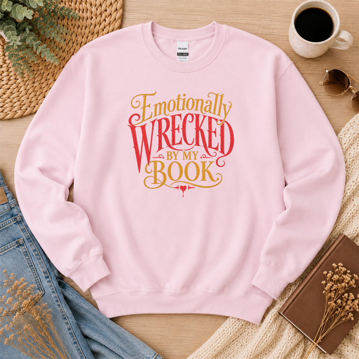 Light pink crewneck sweatshirt laid flat with ornate red and gold lettering and the words “Emotionally Wrecked by My Book.”