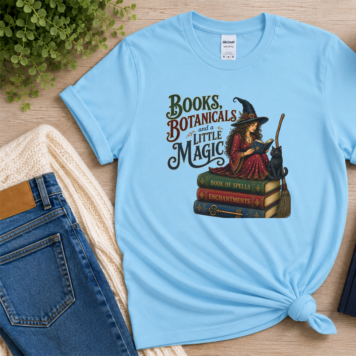 Light blue T-shirt laid flat with a witch reading on stacked spell books beside a black cat and broom and the words “Books, Botanicals and a Little Magic.”