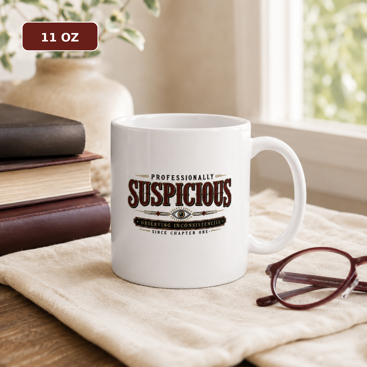11 oz white ceramic mug reading “Professionally Suspicious—Observing Inconsistencies Since Chapter One,” on a cloth-covered table beside stacked books and glasses.
