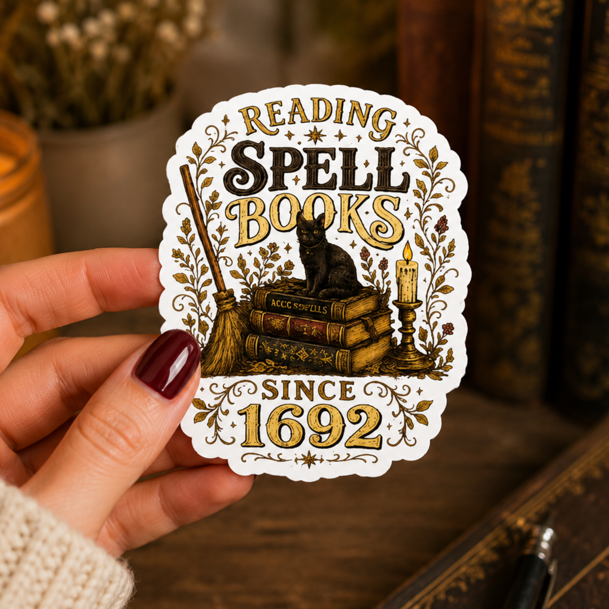 Hand holding a white-bordered sticker reading “Reading Spell Books Since 1692,” featuring black and gold lettering, a black cat on antique books, a broom, candle, and botanical flourishes.