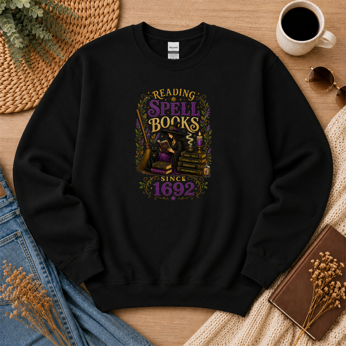 Black crewneck sweatshirt laid flat with a witch reading beside stacked books and candles and the words “Reading Spell Books Since 1692.”