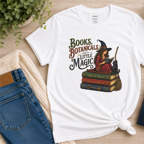 White T-shirt laid flat with a witch reading on stacked spell books beside a black cat and broom and the words “Books, Botanicals and a Little Magic.”