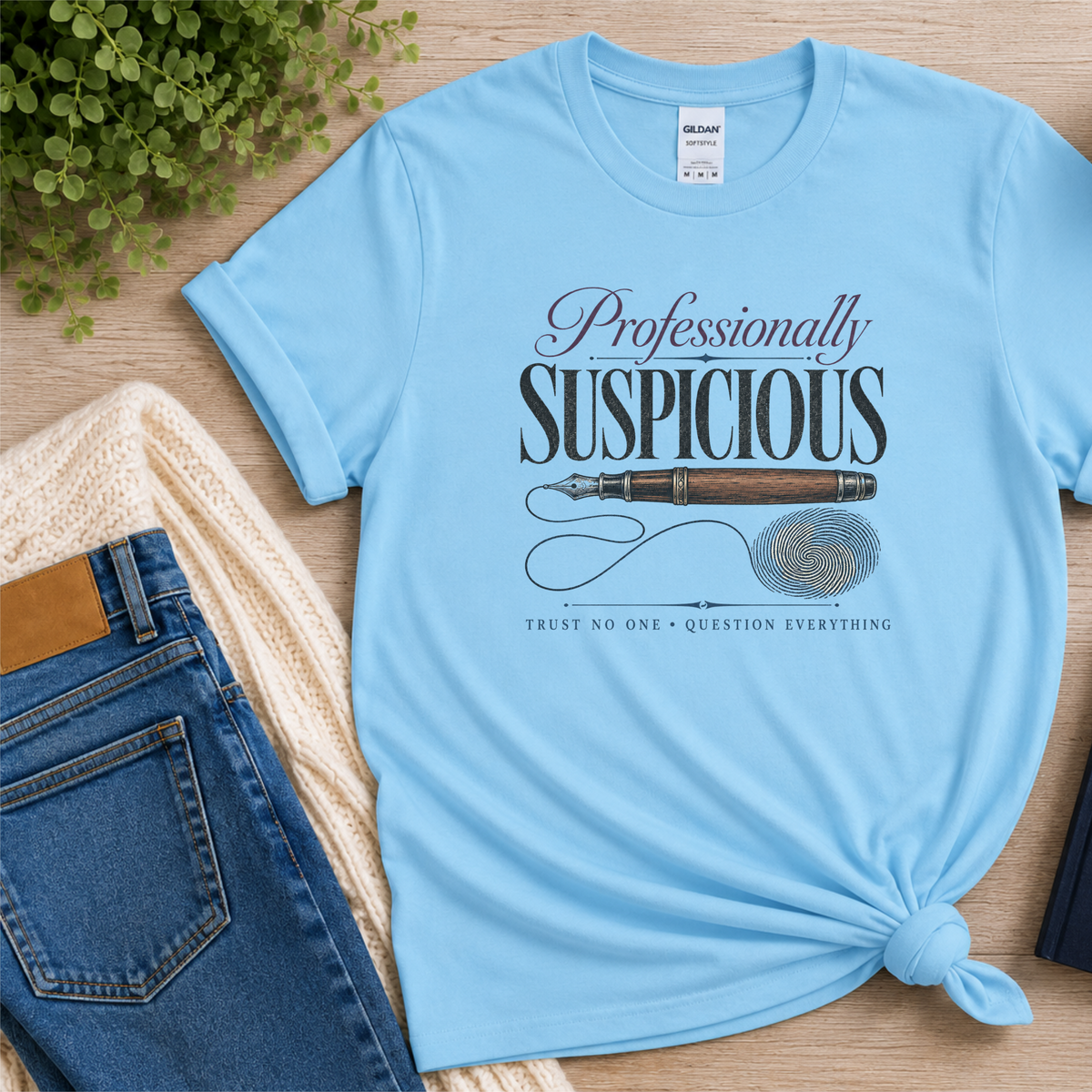 Light blue T-shirt laid flat with a fountain pen and fingerprint and the words “Professionally Suspicious. Trust No One. Question Everything.”