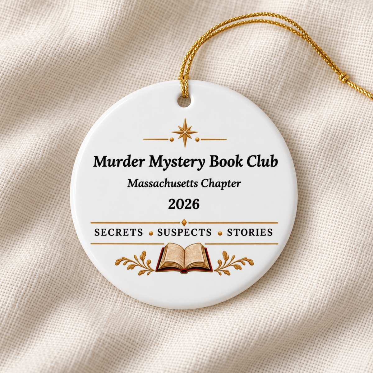Close-up of a white round ornament on cream fabric, with gold string, star and open book; reads “Murder Mystery Book Club,” “Massachusetts Chapter,” “2026” and “Secrets • Suspects • Stories.”