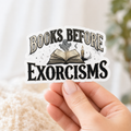 Hand holding a white-bordered sticker reading “Books Before Exorcisms,” featuring black lettering, an open book, gray ghost, candle, and cobweb.